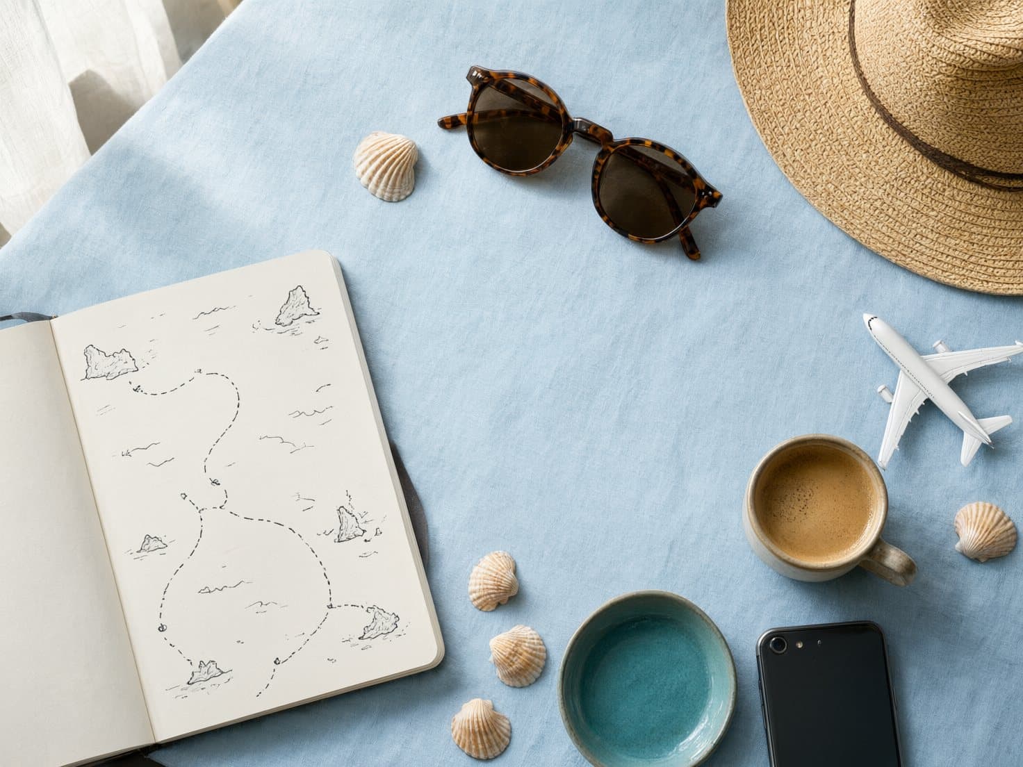 Caderno com um roteiro desenhado, óculos de sol, chapéu de palha e um aviãozinho sobre a mesa